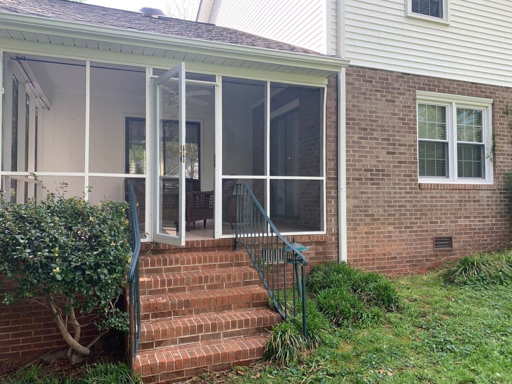 Screened porch entrance with brick steps and greenery outside a residential home.