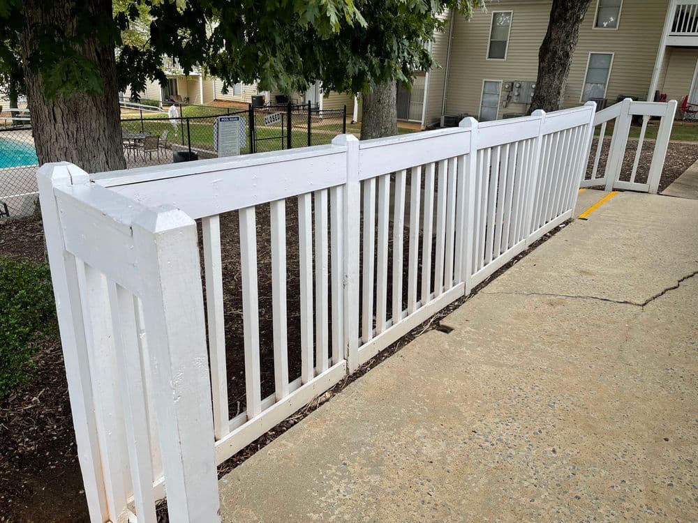 White wooden fence along a paved walkway near a swimming pool at an apartment complex.