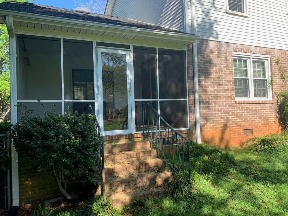 Screened porch with steps at a residential home, surrounded by green grass and shrubbery.