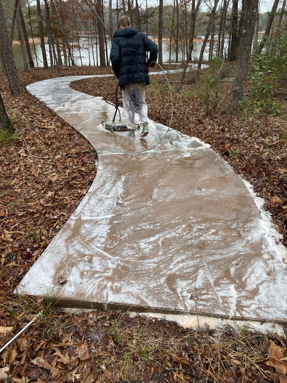 Person smoothing wet concrete on a winding path surrounded by trees and fallen leaves.