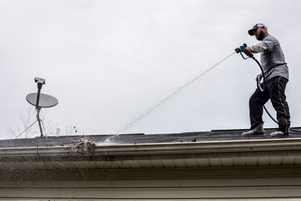 Man cleaning a roof gutter with a pressure washer, removing debris and leaves.