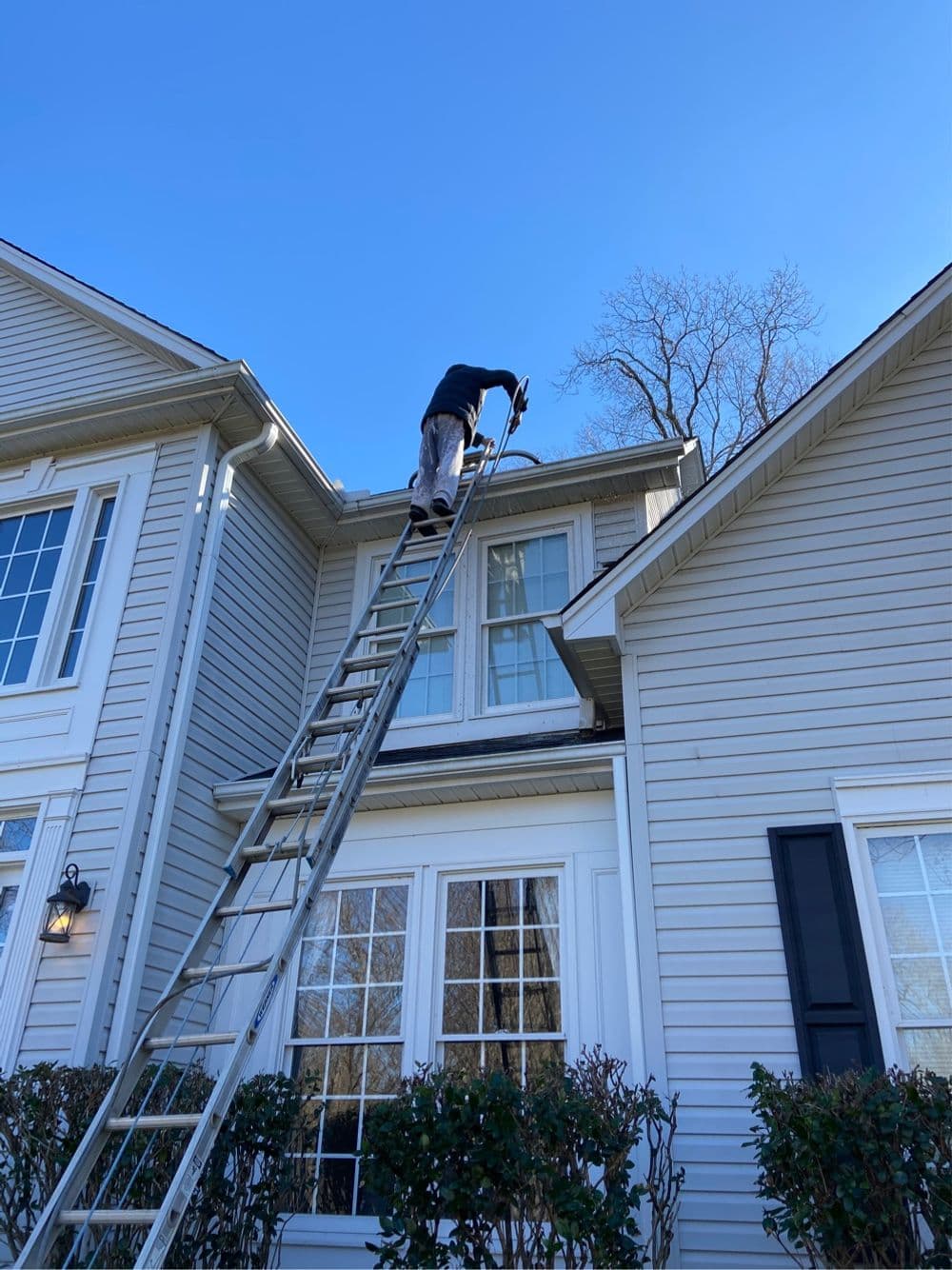 Man on ladder cleaning gutters of a house under a clear blue sky.
