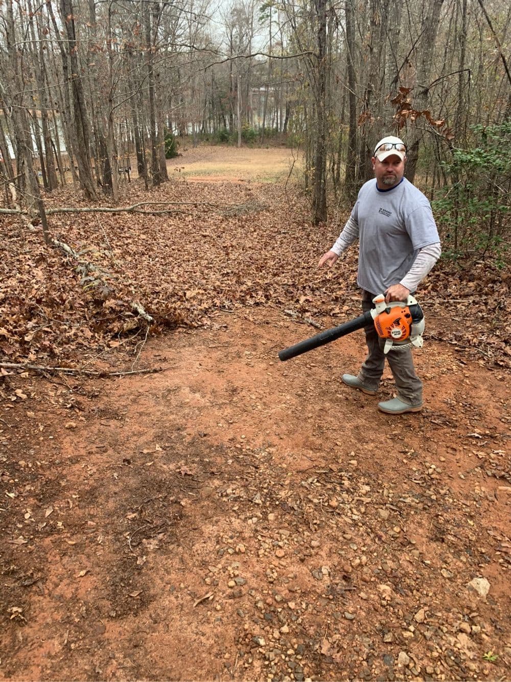 Man using a leaf blower on a wooded trail covered with autumn leaves.