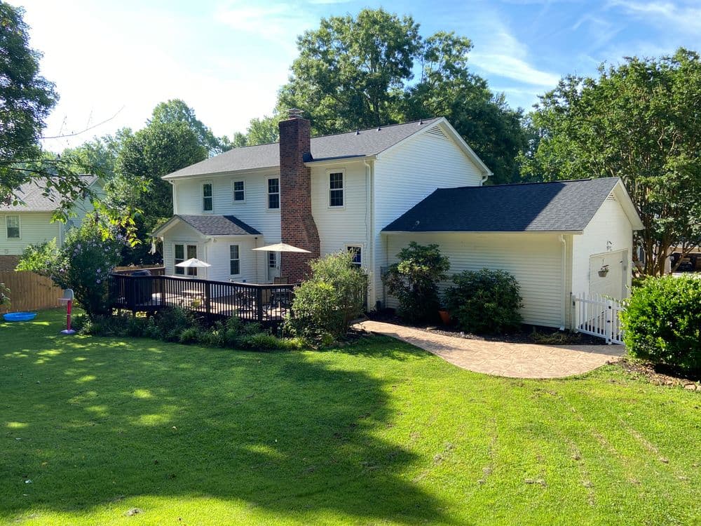 Two-story white house with a deck, surrounded by green lawn and trees on a sunny day.