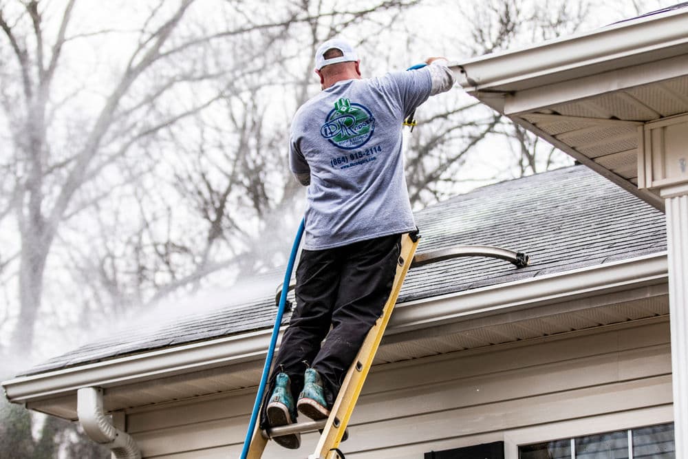 Worker cleaning gutters from a ladder, wearing a grey shirt and safety gear.
