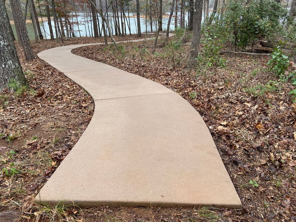 Curved concrete path through wooded area by a lake with autumn leaves scattered along the ground.