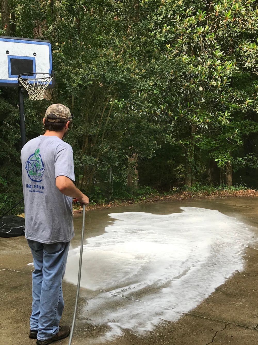 Man using a hose to wash a basketball court outdoors, surrounded by trees.