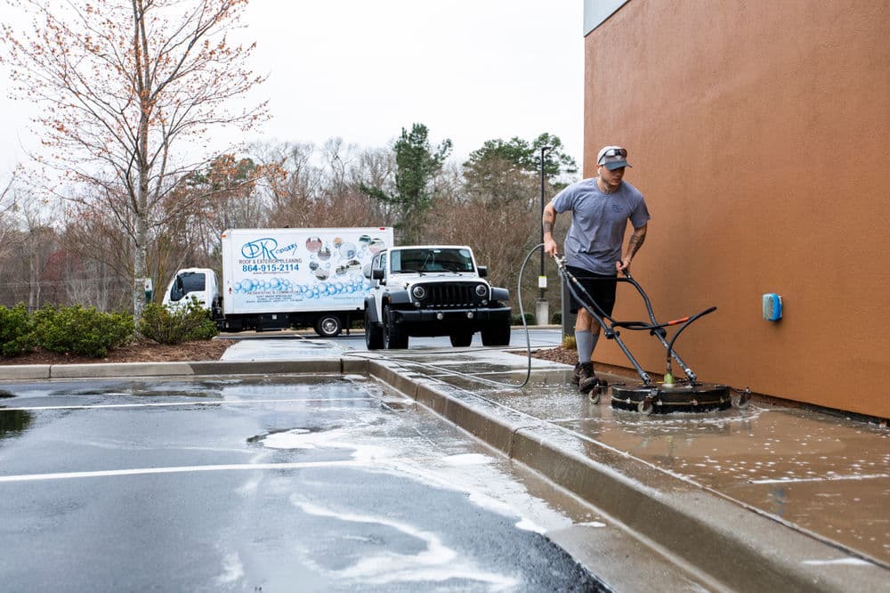 Man power washing a sidewalk with a truck and trailer in the background on a cloudy day.