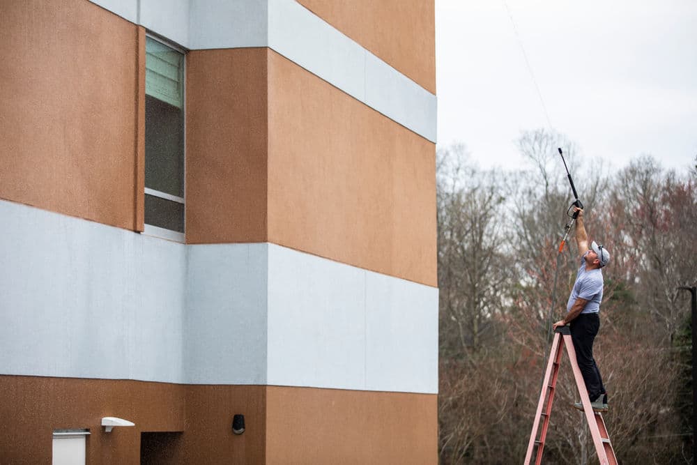 Worker on ladder using a pole to clean a window on a tall building exterior.