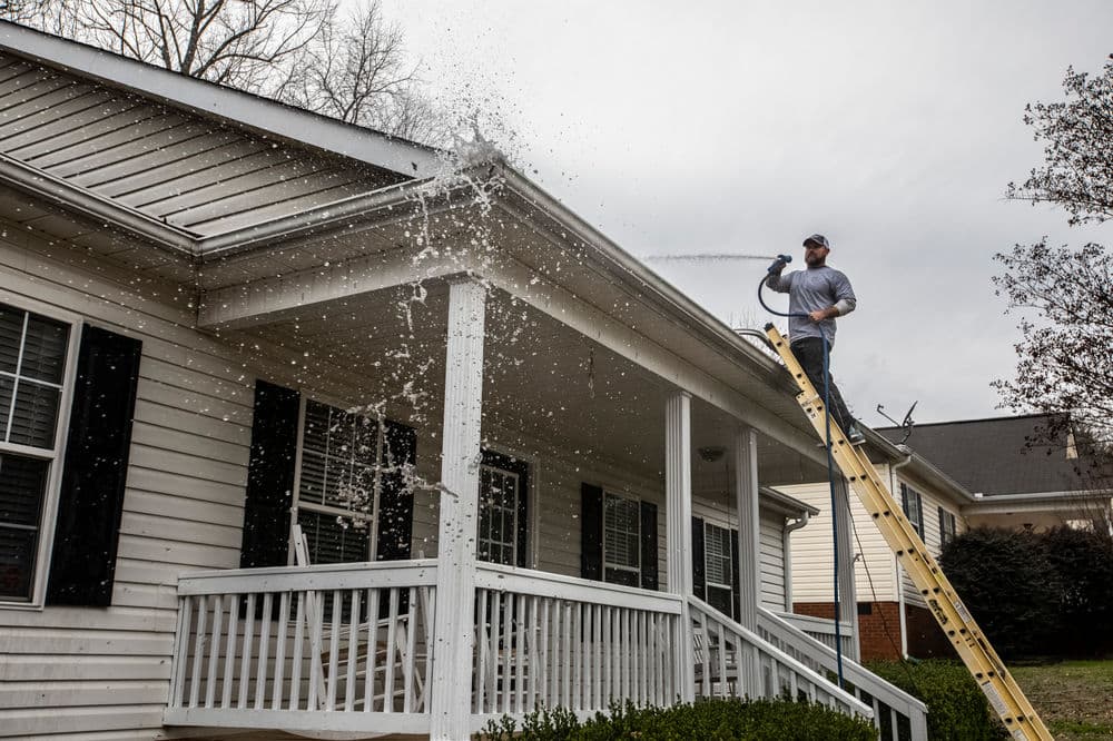Man cleaning roof gutters with pressure washer from a ladder on a residential home.