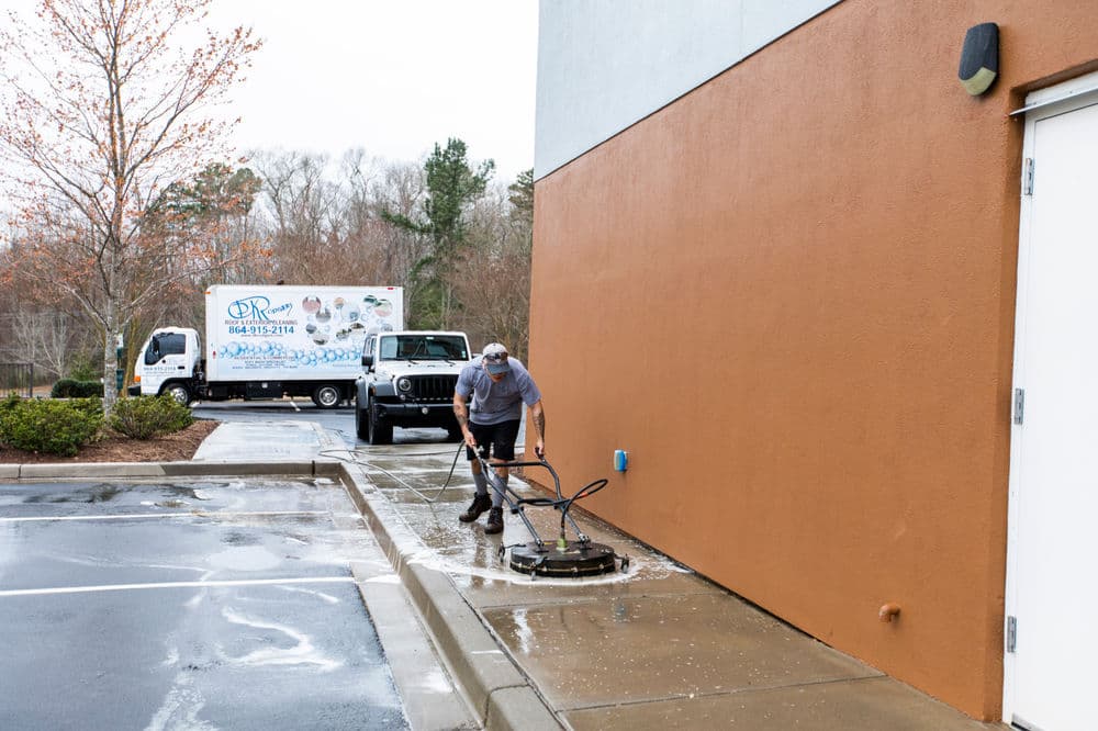 Man pressure washing a sidewalk next to a building with delivery trucks in the background.