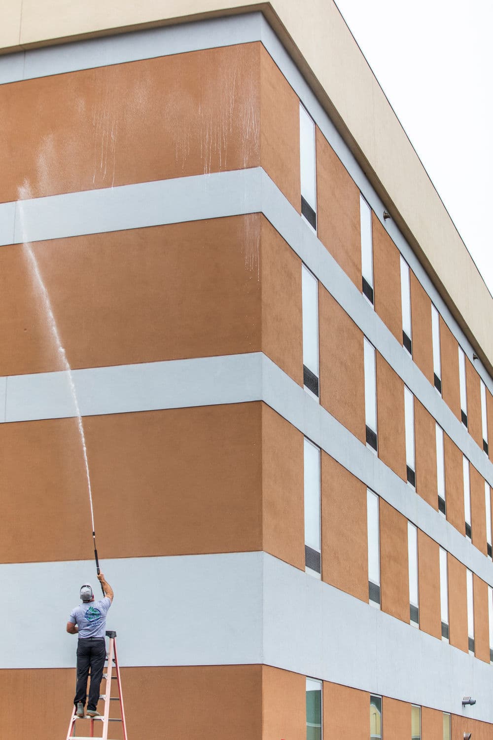 Worker using a pressure washer on a tall building's exterior from a ladder.