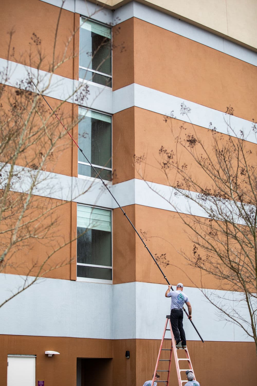 Worker on ladder painting exterior of a multi-story building with a long pole.