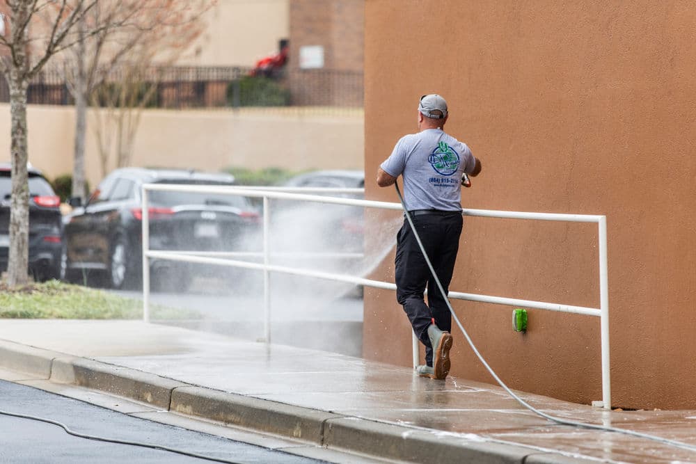 Man pressure washing a sidewalk beside a building with a hose and water spray.