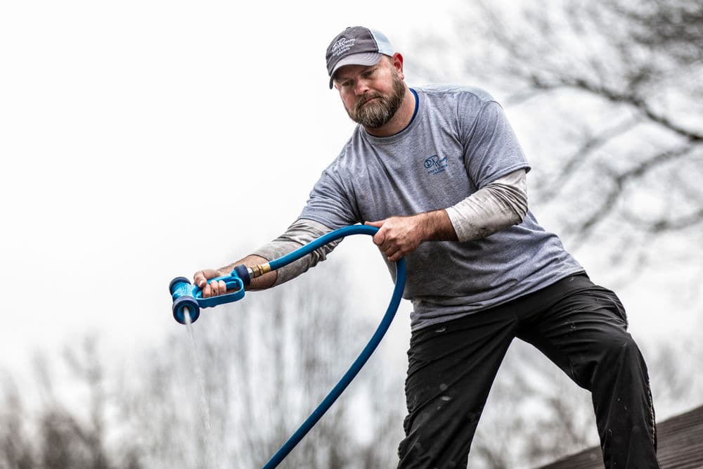 Man using a garden hose, spraying water outdoors, wearing a gray shirt and cap.