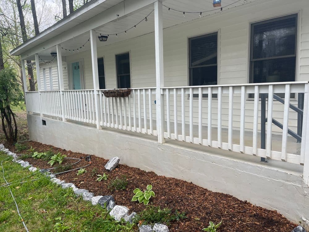 Front porch of a house featuring a white railing, garden beds, and hanging string lights.