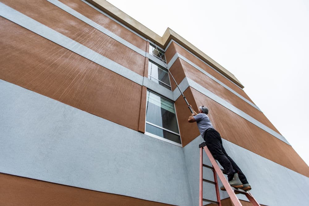 Man painting a building's exterior with a roller while standing on a ladder.