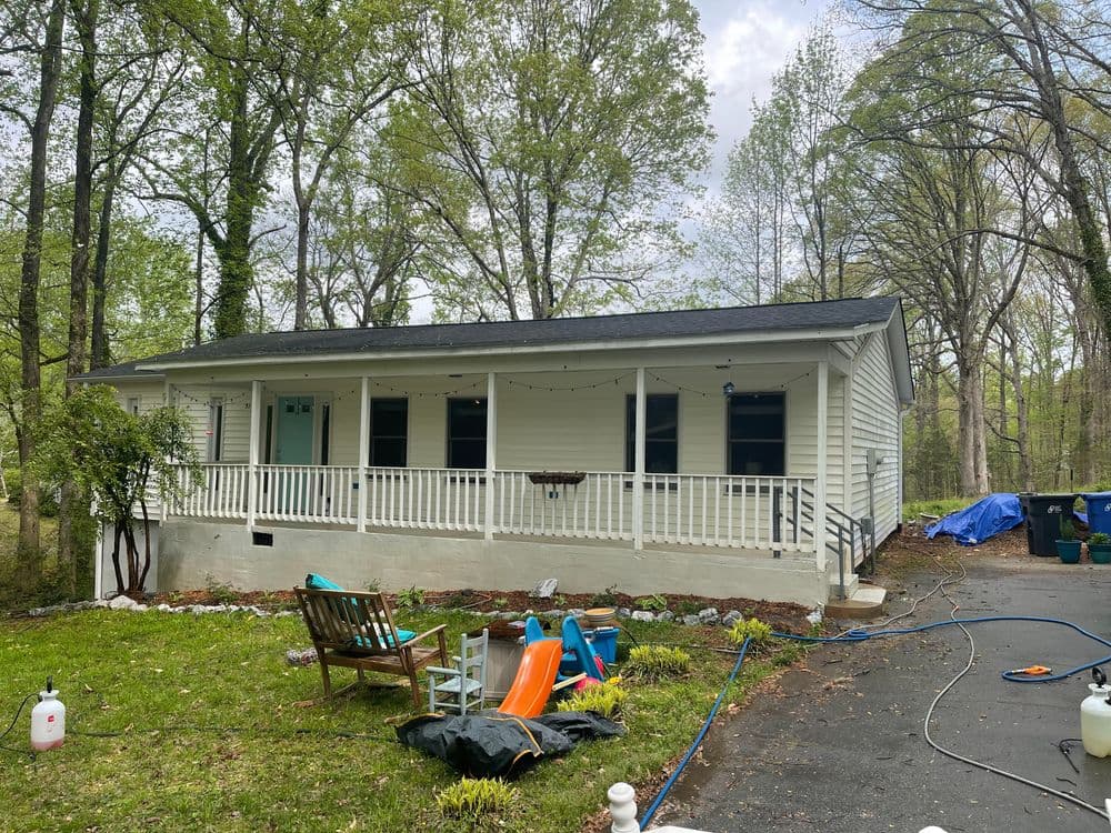 White house with porch surrounded by greenery and lawn furniture in a wooded area.