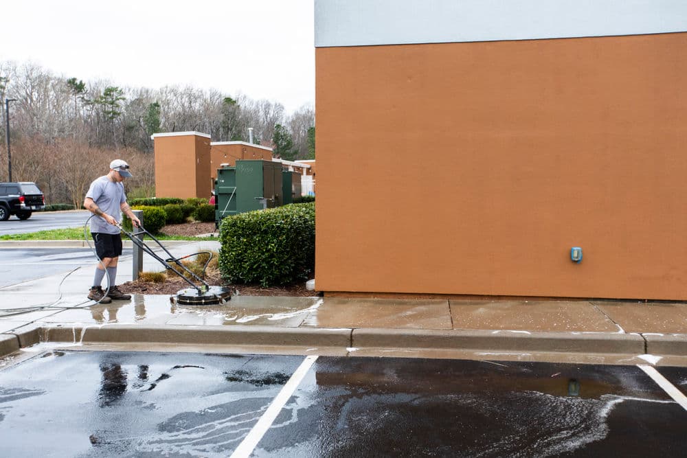 Man pressure washing a sidewalk next to a brown building on a rainy day.