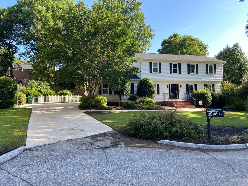 Two-story white house with green shutters, large yard, and paved driveway surrounded by trees.
