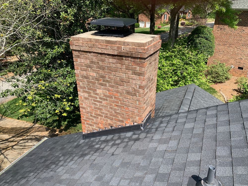 Chimney on a shingled roof with surrounding trees and houses in the background.