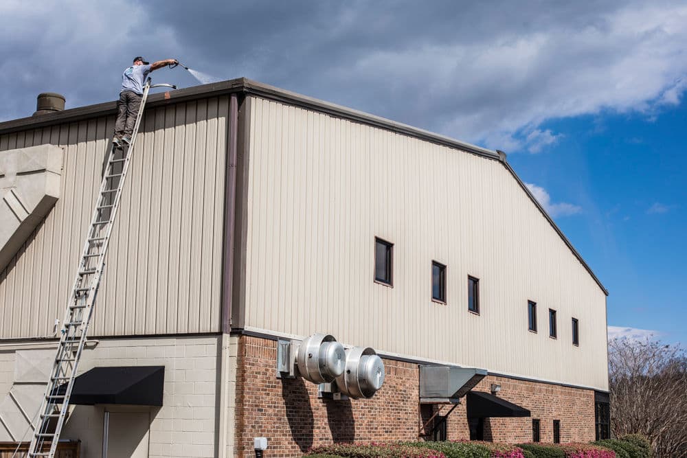Worker on ladder performing maintenance on a building roof under a cloudy sky.