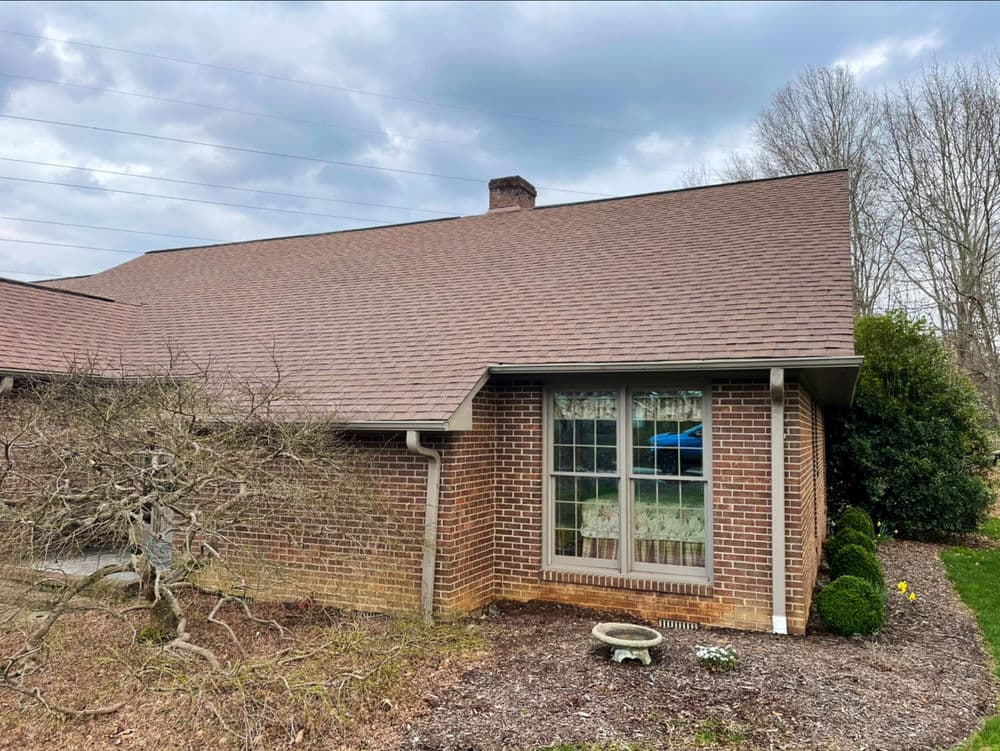 Brick home exterior with a sloped roof, large window, and landscaped yard under cloudy sky.