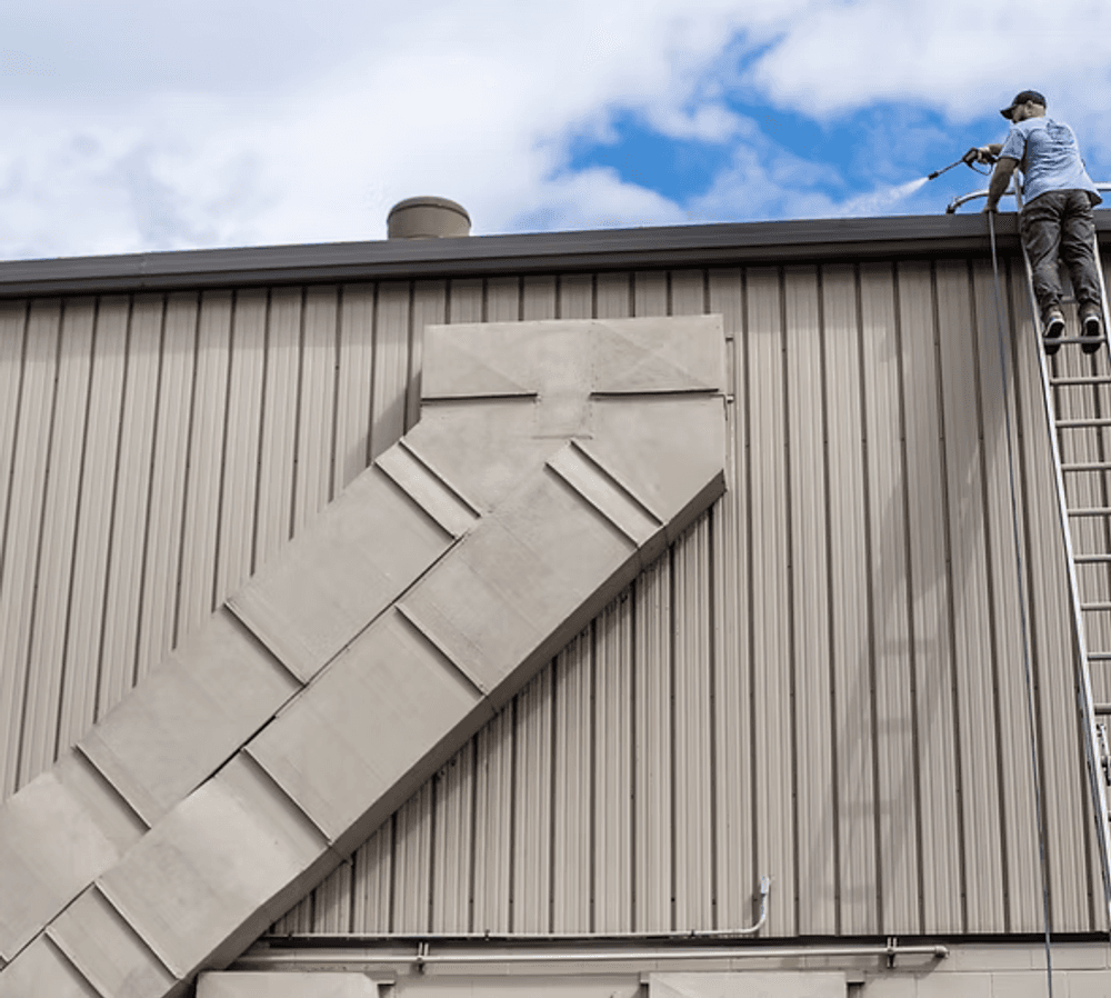 Worker on a ladder inspecting a metal building's roof under a blue sky.
