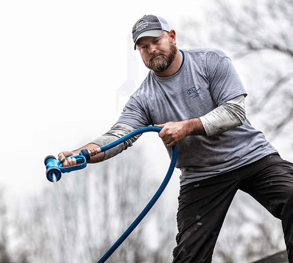 Man using a blue garden hose while standing outdoors, wearing a gray shirt and cap.